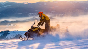 MAN RIDING A SNOWMOBILE IN THE MOUNTAINS WITH THE SUN SETTING BEHIND HIM